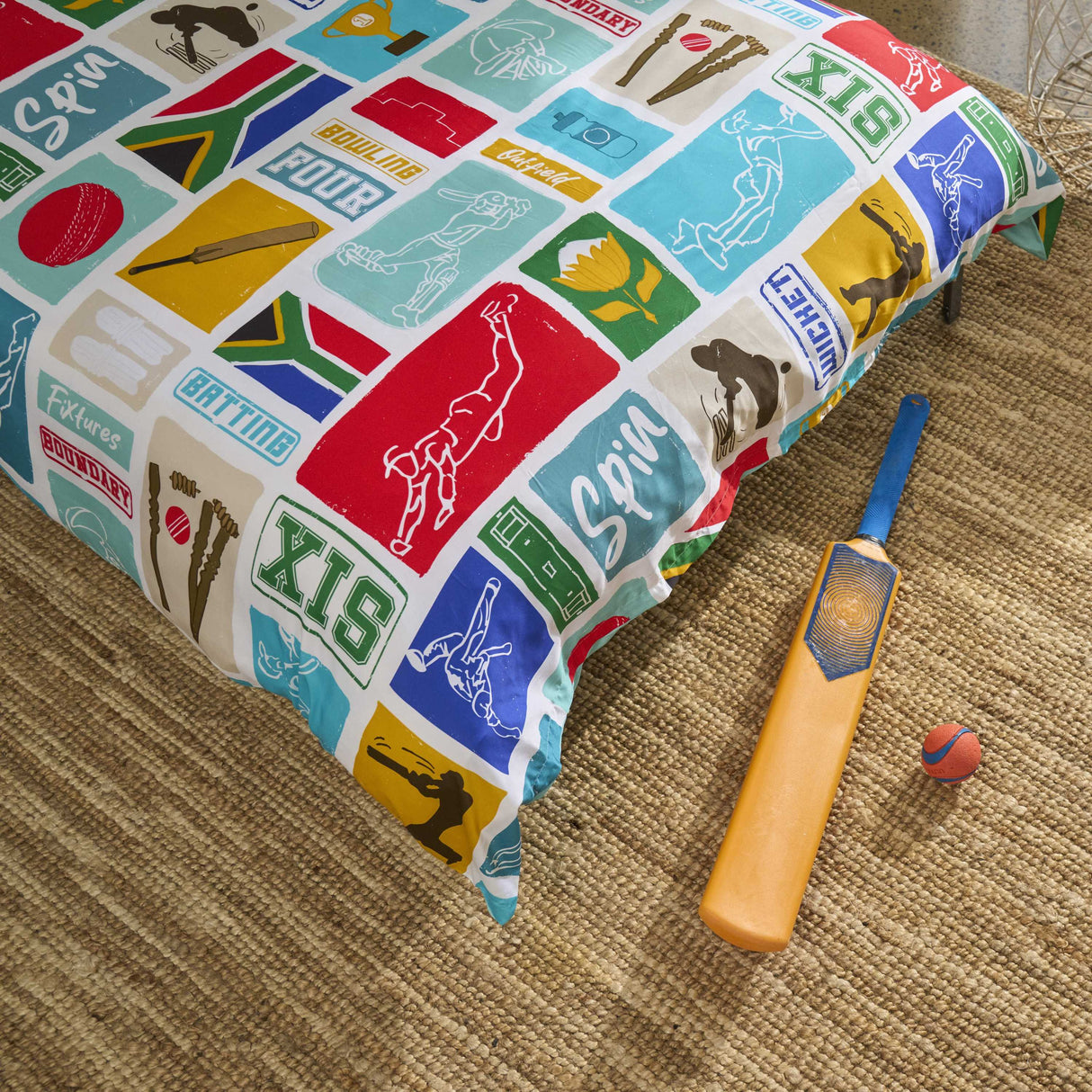 A bed made with a South African cricket-themed printed  duvet cover, styled with a wooden bat and a small ball on a woven jute rug.