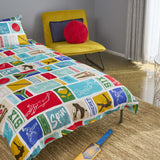 A bed dressed in a colourful South African Cricket-themed duvet cover and pillowcase, shown in a styled bedroom with a yellow chair, red cushion, window, and woven rug. Jo's Dreamland family designed organic kids bedding South Africa.