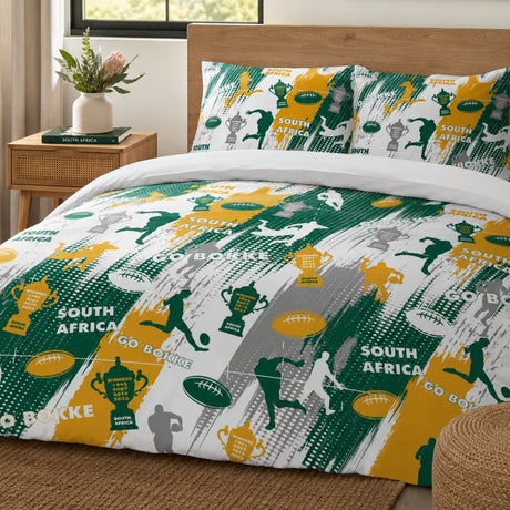 A bed dressed in a South Africa rugby themed duvet cover and matching pillowcases, styled in a bright bedroom with a wooden headboard, bedside table, plant, and woven rug.