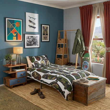 A styled bedroom scene featuring a bed dressed in a white duvet cover and pillowcase set printed with a green camouflage-style skateboard pattern. The room has a blue wall, framed art, a wooden nightstand, a rustic chest, and a window with red curtains.