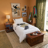 A styled kids skateboarding themed bedroom scene with a bed dressed in white bedding featuring a large black and tan skateboarder print. The room has warm orange walls, green curtains, a wooden nightstand with a lamp, framed wall art, a coat rack with colorful garments, and a dark wood chest at the foot of the bed.