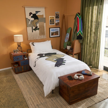 A styled kids skateboarding themed bedroom scene with a bed dressed in white bedding featuring a large black and tan skateboarder print. The room has warm orange walls, green curtains, a wooden nightstand with a lamp, framed wall art, a coat rack with colorful garments, and a dark wood chest at the foot of the bed.