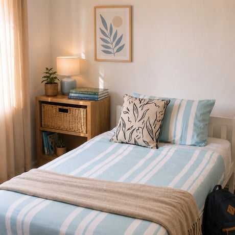 A neatly made bed in a bright bedroom setting, featuring a light blue and white striped duvet cover and matching pillow shams, a beige throw blanket, a patterned accent pillow, and a wooden nightstand with a lamp and books.