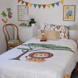 A children's bedroom scene showing a bed made up with a white duvet cover printed with a cartoon lion and green tree, plus matching animal-print pillows and a giraffe plush toy. The room includes wall decals, framed animal artwork, a small cabinet, chair, and colorful bunting.