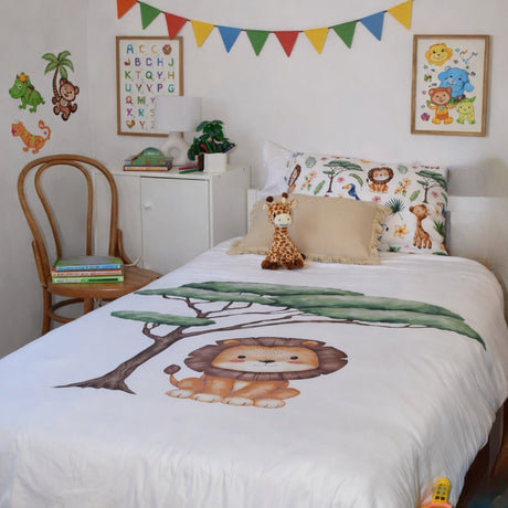 A children's bedroom scene showing a bed made up with a white duvet cover printed with a cartoon lion and green tree, plus matching animal-print pillows and a giraffe plush toy. The room includes wall decals, framed animal artwork, a small cabinet, chair, and colorful bunting.