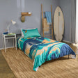 Bedroom with a bed featuring a colourful surfing themed duvet cover, a surfboard against the wall, and a nightstand with a lamp.