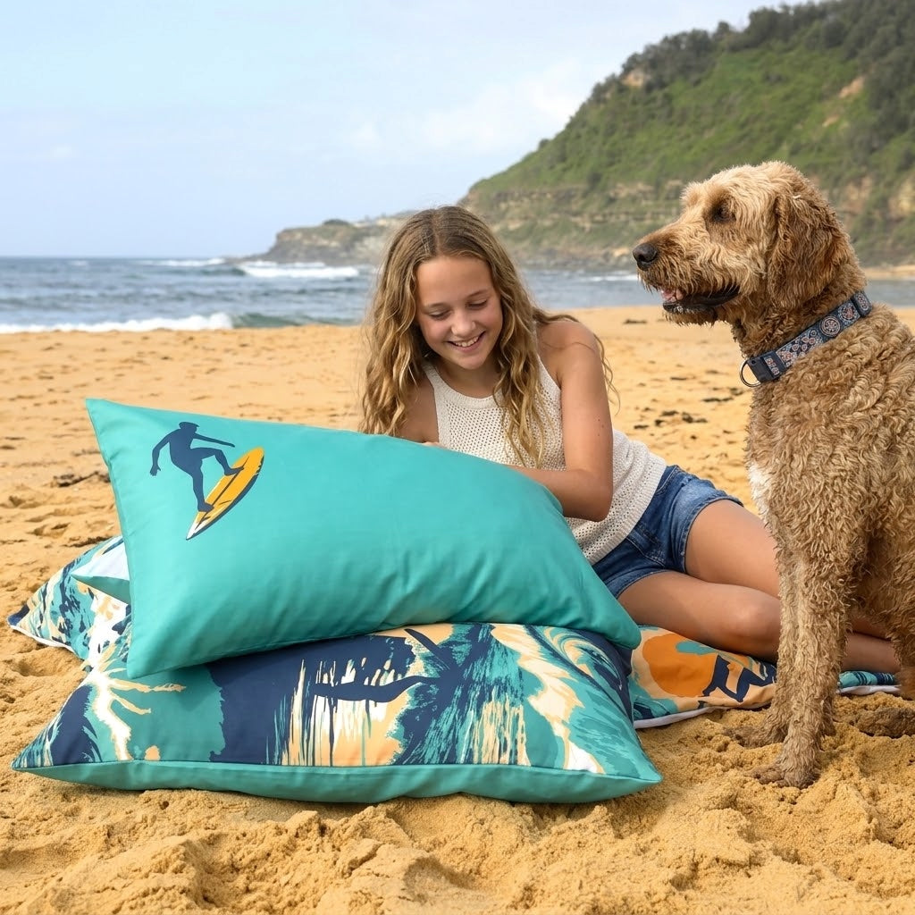 A girl and her dog relax on a beach with a teal pillow with a surfer in the corner. The duvet cover feature a turquoise and navy tropical beach print with a surfboard graphic on the top pillow.