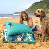 A girl and her dog relax on a beach with a teal pillow with a surfer in the corner. The duvet cover feature a turquoise and navy tropical beach print with a surfboard graphic on the top pillow.