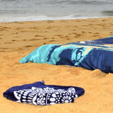A navy blue blanket with a white bohemian-style pattern is folded on a sandy beach, with a larger matching blanket in a turquoise and navy print lying further back near the shoreline.