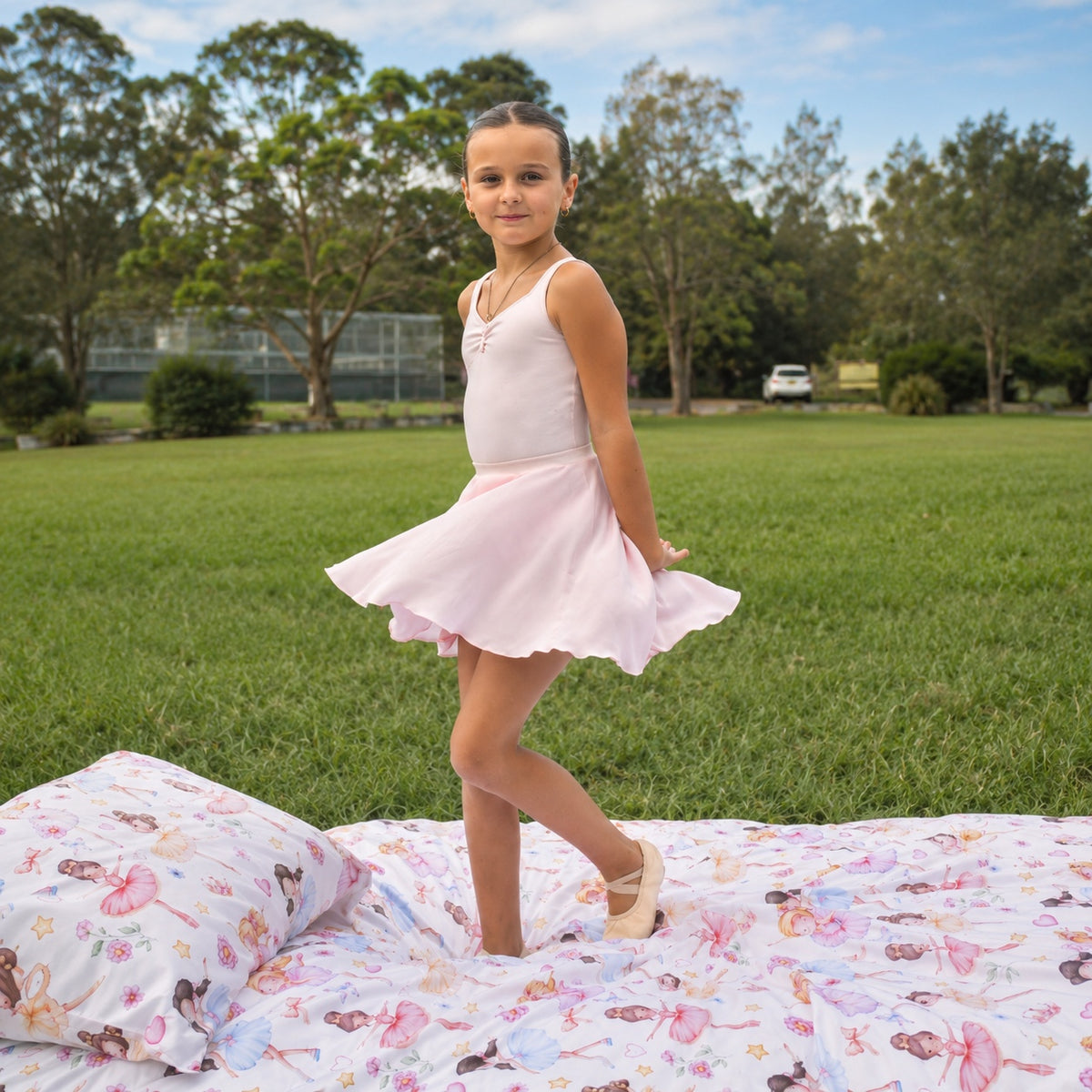 A young girl in a light pink sleeveless dance dress stands on a colorful ballet-print duvet cover set on a grassy lawn, with trees and a park in the background.