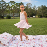 A young girl in a light pink sleeveless dance dress stands on a colorful ballet-print duvet cover set on a grassy lawn, with trees and a park in the background.