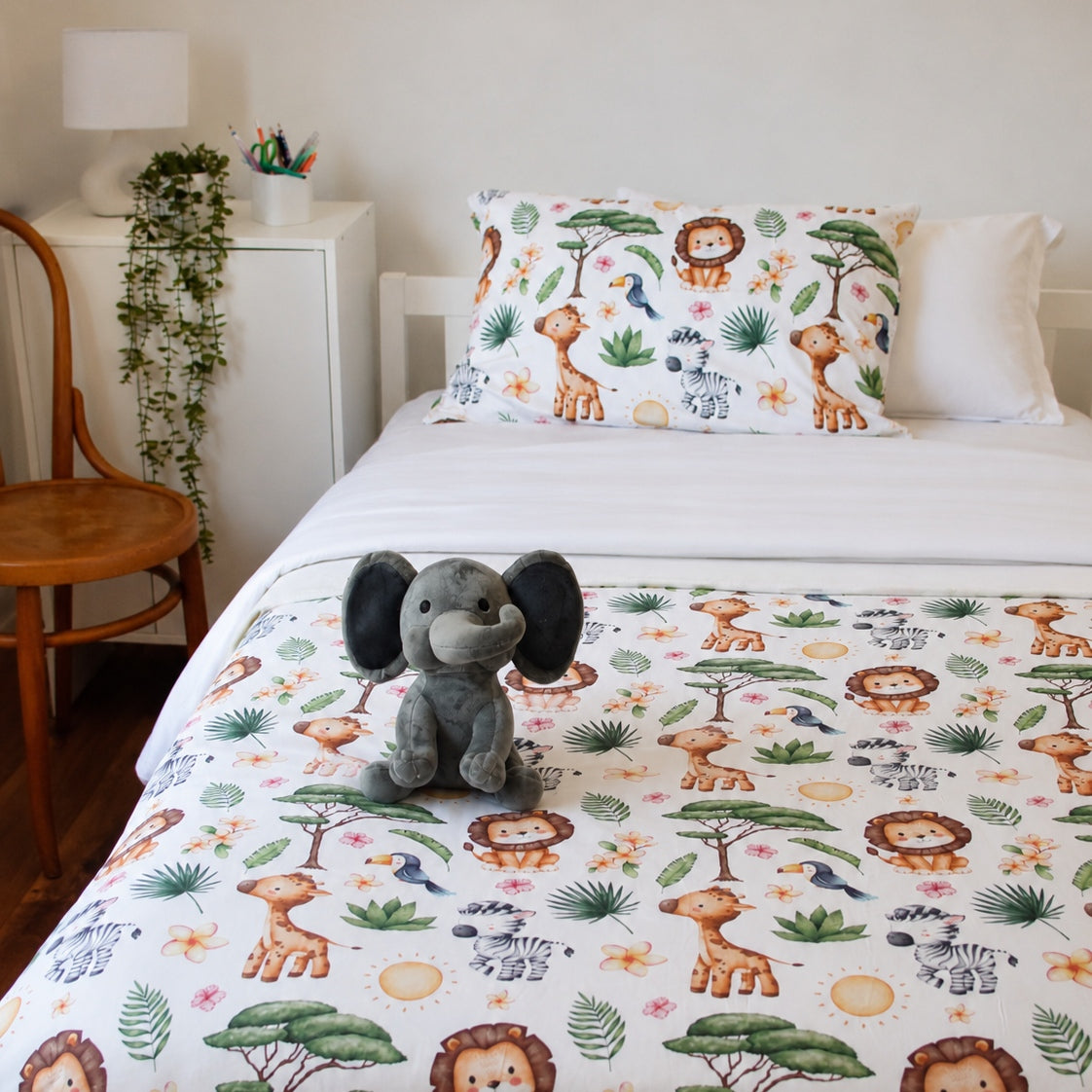 A children's bed styled in a bedroom setting, featuring a white duvet cover and matching pillowcase printed with a colorful safari animal pattern including lions, giraffes, zebras, and a gray elephant plush toy sitting on the bed.