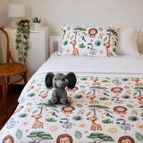 A children's bed styled in a bedroom setting, featuring a white duvet cover and matching pillowcase printed with a colorful safari animal pattern including lions, giraffes, zebras, and a gray elephant plush toy sitting on the bed.