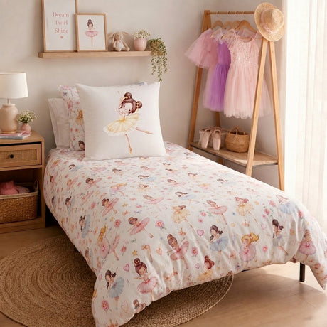 A children's bed dressed in a white ballerina-themed bedding set with a matchingcontinental pillow, styled in a soft pink bedroom with a wooden floor, bedside table, lamp, wall shelf, and a clothing rack holding pink and purple dresses.